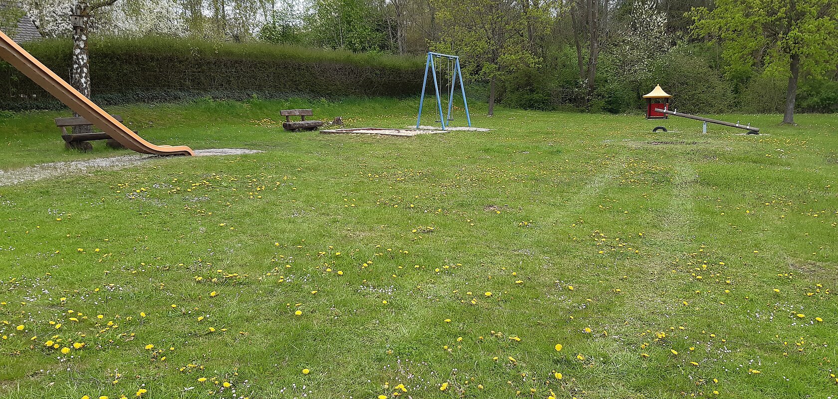 Spielplatz mit Rutsche, Schaukel, Wippe, zwei Bänken und Wiese mit gelben Blumen. Im Hintergrund sind Bäume bei bewölktem Himmel.