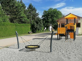 Spielplatz mit Schottersteinen einer schwarz-gelber Nestschaukel und schwarz-orangem Klettergerüst vor einem unverputzten Haus und Bäumen links davon, bei sonnigem Himmel.