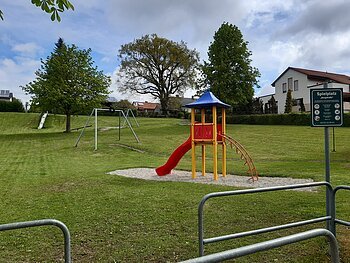 Spielplatz mit Rutsche, Klettergerüst, Seilbahn, Schaukel und Hinweisschild auf einer Wiese vor Wohnhäusern bei bewölktem Himmel.