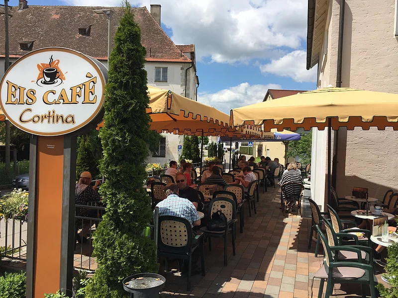 Eiscafé Cortina Außenbereich eines Eiscafés mit gelben Sonnenschirmen, Tischen und Stühlen, Gäste sitzen und essen.
