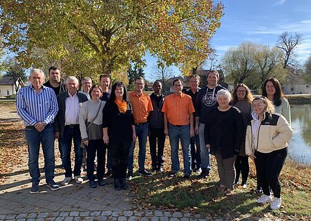 Gruppenfoto von 16 Personen im Freien vor einem großen Baum und einem Teich rechts im Bild, an einem sonnigen Herbsttag.