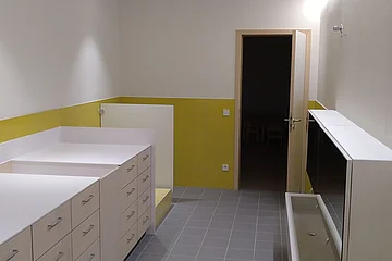 Sanitärbereich im Kindergarten Monheim: ein Raum mit weißen breiten Schränken mit Ablagefläche, gelb-grünen Wandfliesen bis zur Hälfte der Wände, grauem Boden und eine offener Tür zum nächsten Raum.