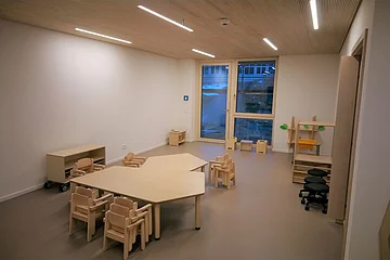 Gruppenraum im städtischen Kindergarten Monheim mit Kindertischen und Stühlen aus Holz, einem Fenster und verschiedenen Regalen.