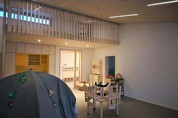 Innenraum eines Kindergartens mit einem grauen Spielzelt links im Bild, kleinen Holztischen und Stühlen, einem Wandschrank aus Holz, sowie einer kleinen Holztreppe in den Spielbereich oberhalb, der mit einem Holzgeländer verkleidet ist.