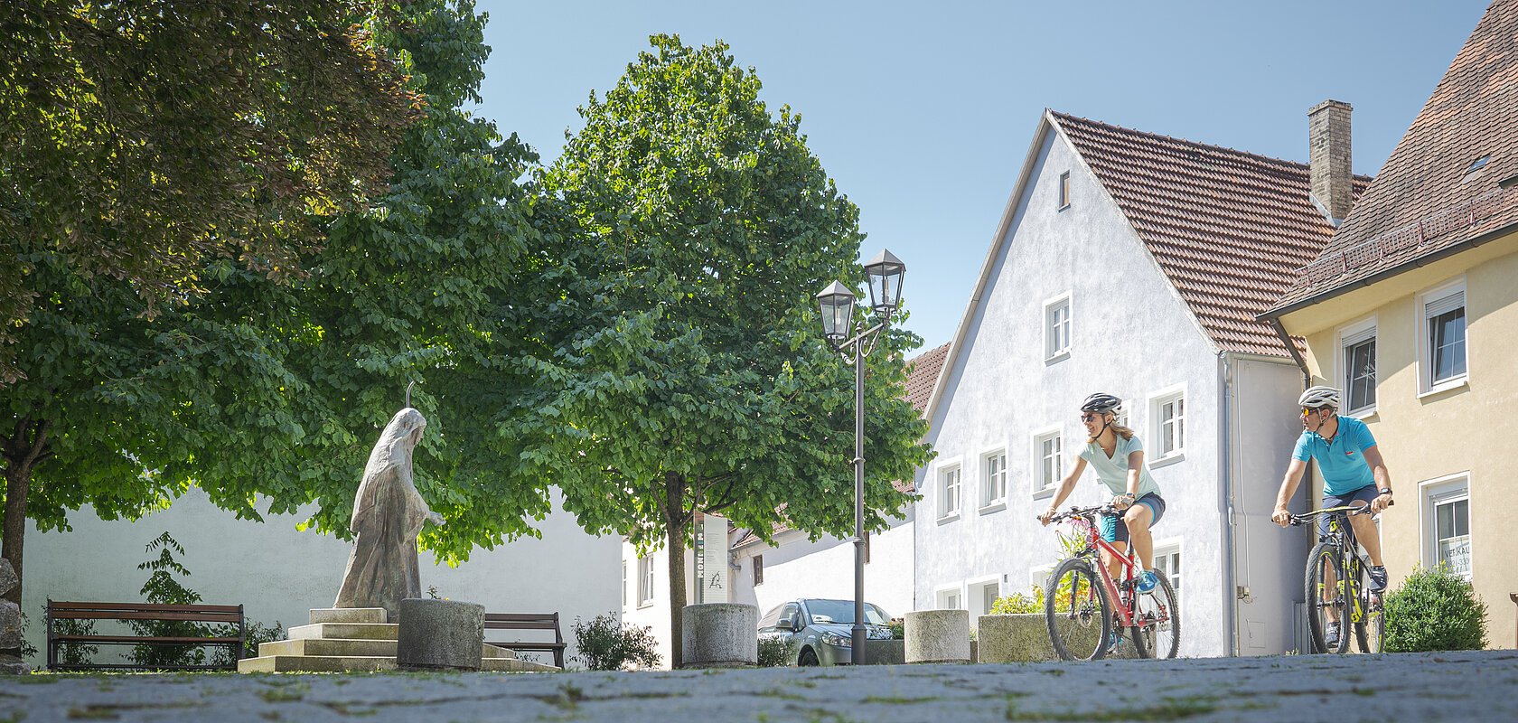 Zwei Radfahrer rechts im Bild fahren auf gepflastertem Platz mit einer Statue und Bänken links im Bild. Bäume und Häuser im Hintergrund.