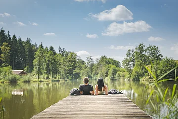 Zwei Personen sitzen auf einem Steg an einem See, umgeben von Bäumen und unter blauem Himmel. Im Hintergrund links im Bild eine kleine Holzhütte zu sehen.
