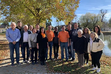 Gruppenfoto von 16 Personen im Freien vor einem großen Baum und einem Teich rechts im Bild, an einem sonnigen Herbsttag.