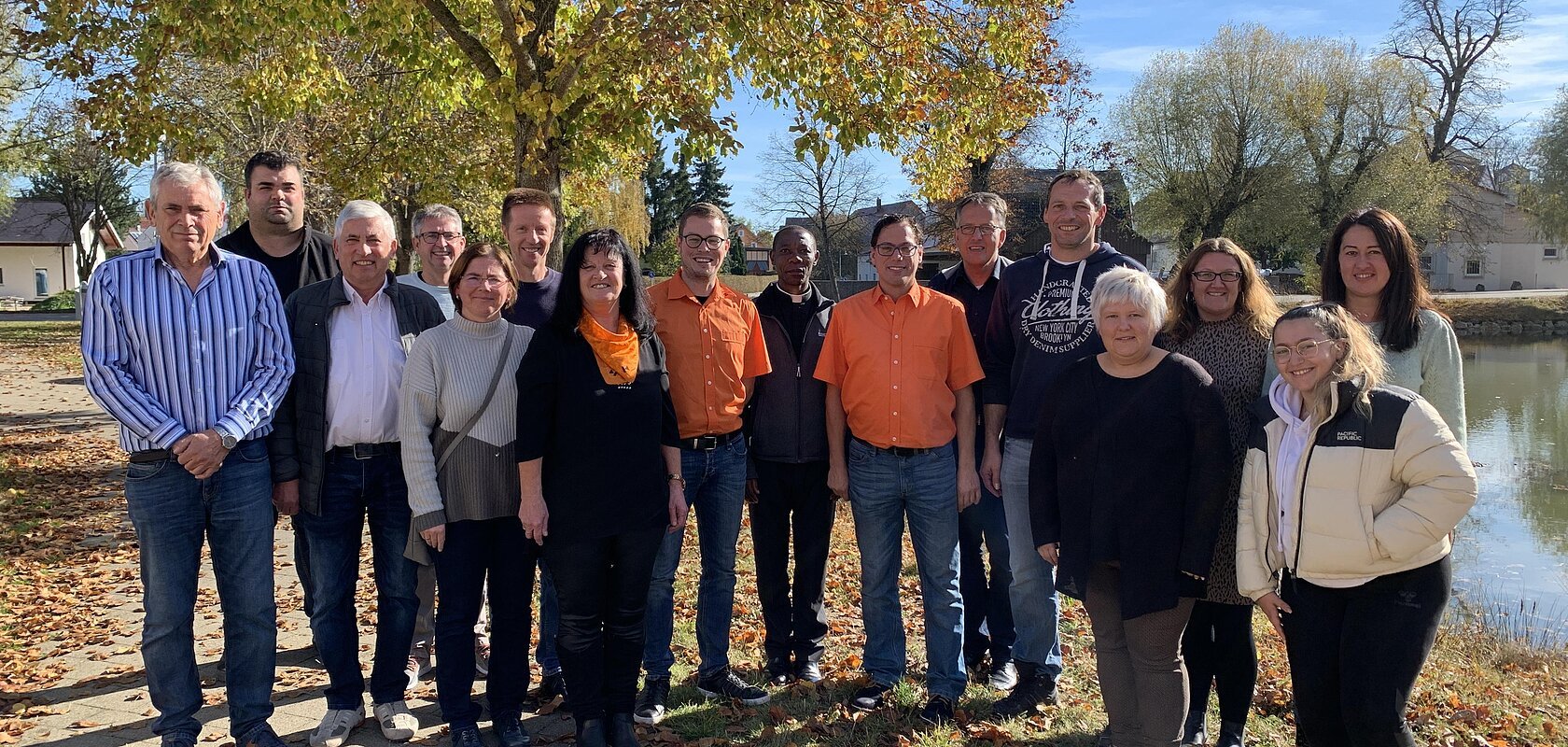 Gruppenfoto von 16 Personen im Freien vor einem großen Baum und einem Teich rechts im Bild, an einem sonnigen Herbsttag.