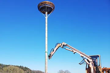In der Mitte des Bildes ein hoher Pfahl mit großem Vogelnestkorb oben auf einem Feld bei Sonnenschein und blauem Himmel. Rechts im Bild steht eine Baumaschine, die den Pfahl aufrichtet.