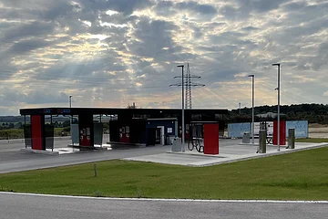 Freie Autowaschanlage mit mehreren Waschplätzen unter bewölktem Himmel und Sonnenstrahlen. Davor eine grüne Rasenfläche.