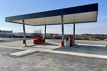 Leere Tankstelle mit zwei roten Zapfsäulen auf einem gepflasterten Platz, unter einem großen Dach bei klarem Himmel.