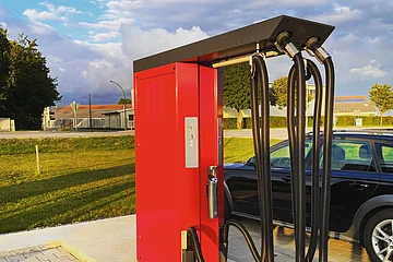 Rote Saug-Station mit schwarzen Schläuchen auf gepflastertem Boden neben schwarzem Auto rechts im Bild, bei Tageslicht. Im Hintergrund grüne Wiese.