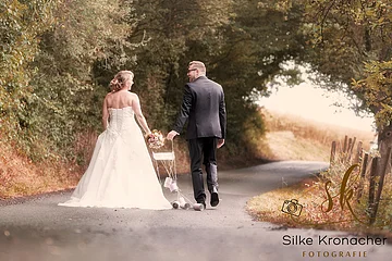 Braut und Bräutigam gehen auf einem gepflastertem Weg, Braut hält in der rechten Hand einen Blumenstrauß, Dosen sind an einem Gebinde befestigt, das von beiden festgehalten Wird. Das Brautpaar schaut sich an. Rechts unten im Bild das Logo der Fotografin Silke Kronacher.