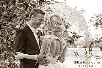 Braun-weiß Foto eines Brautpaares, die Braut hält einen Regenschirm mit Spitze, sie umarmen sich im Freien, unscharfer Hintergrund. Rechts unten im Bild das Logo der Fotografin Silke Kronacher.