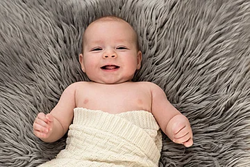 Nackiges Baby liegt auf grauem Fell und ist in eine helle Decke eingewickelt, lächelt in die Kamera. Rechts unten im Bild das Logo der Fotografin Silke Kronacher.