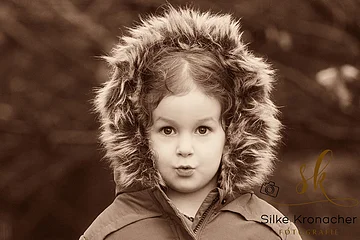 Kind mit Kapuze mit Fellrand blickt in die Kamera, sepiafarbenes Foto, unscharfer Hintergrund. Rechts unten im Bild das Logo der Fotografin Silke Kronacher.