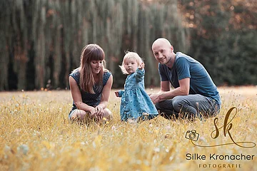 Drei Personen, zwei Erwachsene und dazwischen ein Kleinkind alle in blauer Kleidung, sitzen auf einer Wiese mit gelbem Gras, im Hintergrund unscharf Bäume. Rechts unten im Bild das Logo der Fotografin Silke Kronacher.