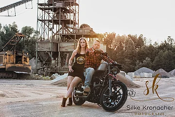 Mann und Frau sitzen auf schwarzem Motorrad vor alter Industrieanlage mit Bagger, Kieshaufen und Bäumen im Hintergrund. Rechts unten im Bild das Logo der Fotografin Silke Kronacher.