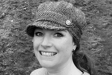 Porträt einer lächelnden Frau mit Fischgrätmuster-Mütze und Pullover vor Grasfläche, Foto in schwarz-weiß.