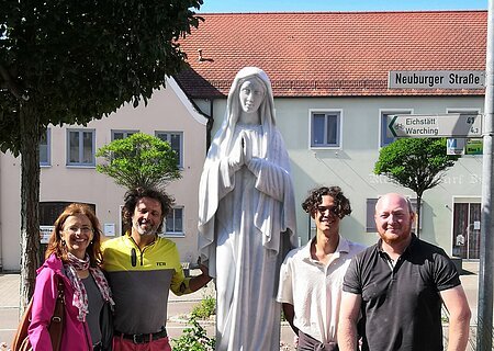 Vier Personen stehen neben einer weißen Marienstatue im Blumenbeet an einer Straße mit Häusern im Hintergrund.