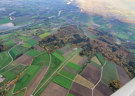 Luftaufnahme von Feldern, Waldflächen und einem Fluss bei Sonnenlicht, mit Dörfern und Straßen.
