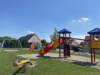 Spielplatz mit Wippe, Rutsche, Klettergerüst, Schaukel und Karussell bei sonnigem Himmel in einem Wohngebiet.