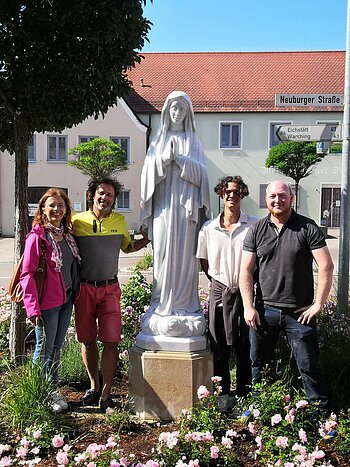 Vier Personen stehen neben einer weißen Marienstatue im Blumenbeet an einer Straße mit Häusern im Hintergrund.