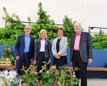 Vier Personen stehen vor einer bepflanzten Mauer mit Rosen und anderen Blumen im Vordergrund.