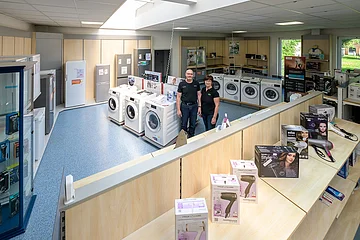 Verkaufsraum mit Haushaltsgeräten, zwei Personen stehen nebeneinander zwischen Waschmaschinen und Kühlschränken. Im Vordergrund Regale mit verschiedenen Ausführungen von Föhnen.