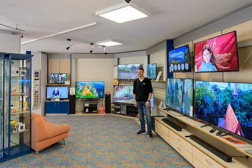 Mann steht in einem Elektronikgeschäft vor mehreren an der Wand montierten Fernsehern mit verschiedenen Bildschirminhalten. Links im Bild ein organgefarbenes Sofa.