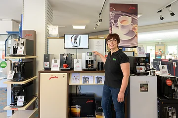 Frau steht in Elektronikgeschäft vor Jura-Kaffeemaschinen und zeigt auf eine Maschine mit Kaffeeauslauf.