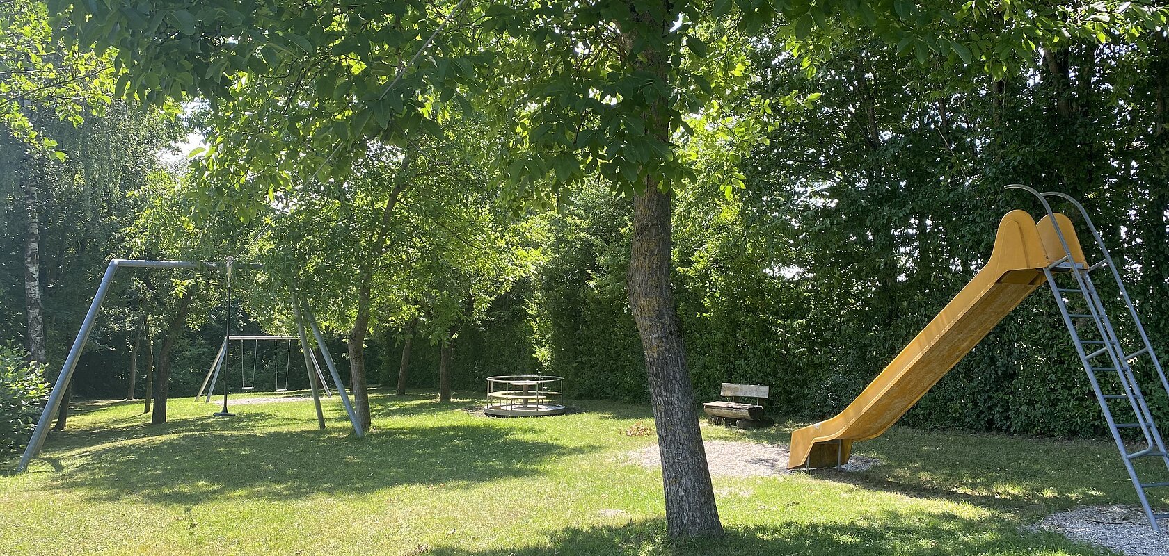 Spielplatz mit Rutsche, Schaukel, Karussell und Bank unter Bäumen auf Grasfläche bei Sonnenschein.