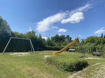 Spielplatz mit gelber Rutsche, zwei Schaukeln und drei Bänken, umgeben von Büschen unter blauem Himmel.
