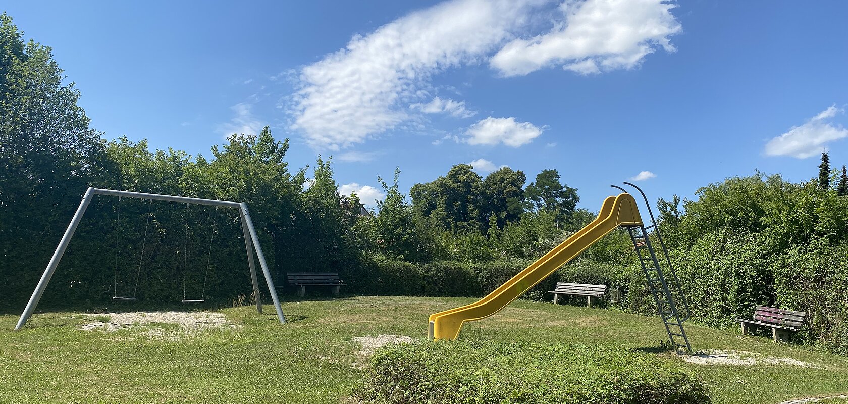 Spielplatz mit gelber Rutsche, zwei Schaukeln und drei Bänken, umgeben von Büschen unter blauem Himmel.