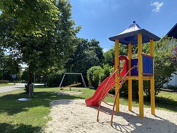 Spielplatz mit Rutsche, Klettergerüst und Schaukel in einem Wohngebiet auf Gras- und Sandfläche bei Sonnenschein.