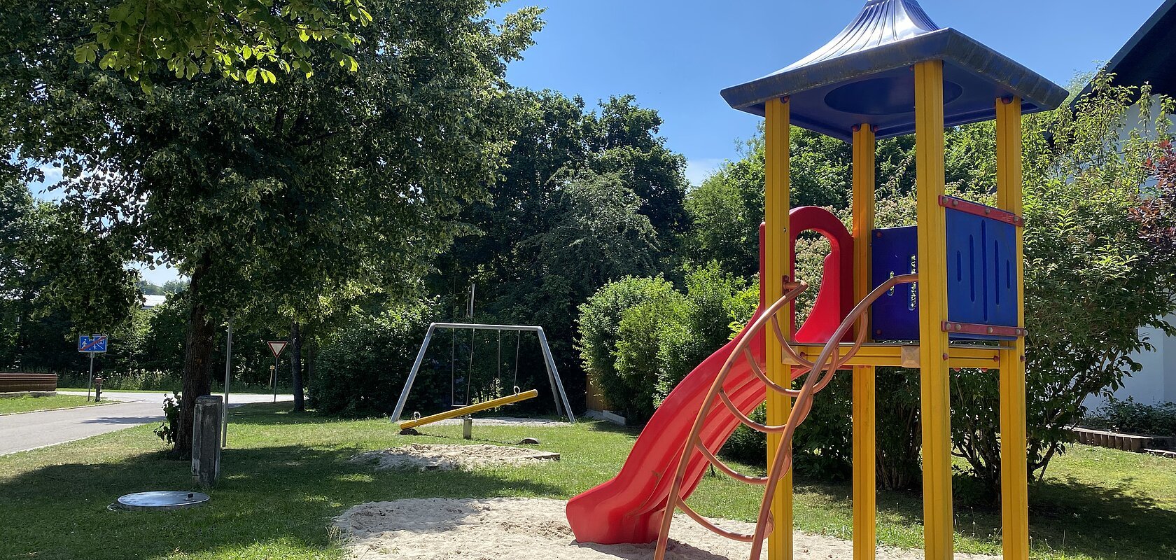 Spielplatz mit Rutsche, Klettergerüst und Schaukel in einem Wohngebiet auf Gras- und Sandfläche bei Sonnenschein.