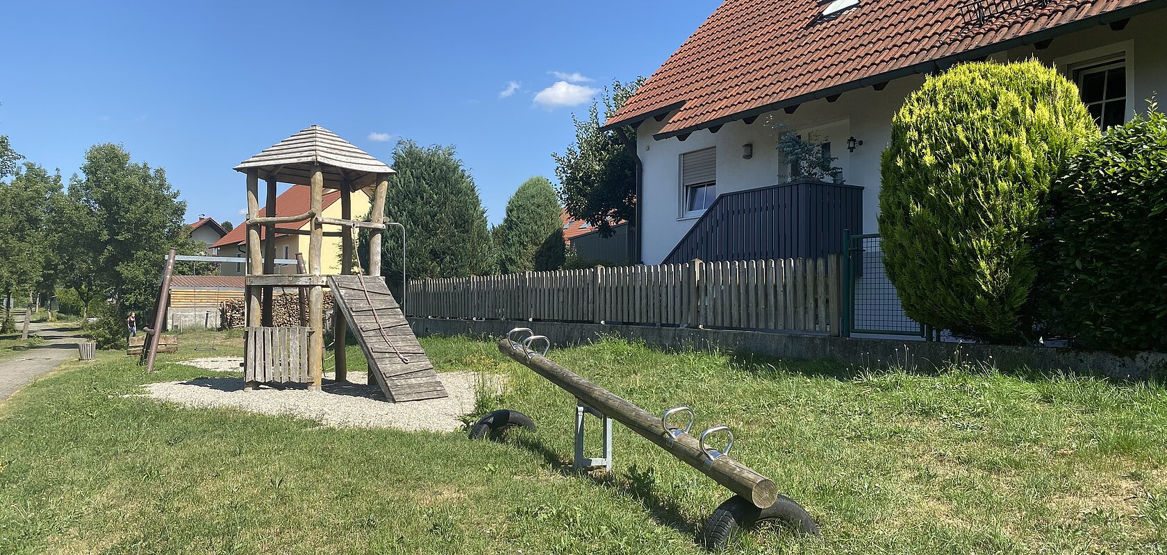 Spielplatz mit Wippe, Schaukel im Hintergrund und Kletterturm auf grünem Rasen, links vor einem Haus mit rotem Dach und Zaun an einem sonnigen Tag.