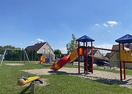 Spielplatz mit Wippe, Rutsche, Klettergerüst, Schaukel und Karussell bei sonnigem Himmel in einem Wohngebiet.