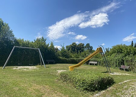 Spielplatz mit gelber Rutsche, zwei Schaukeln und drei Bänken, umgeben von Büschen unter blauem Himmel.