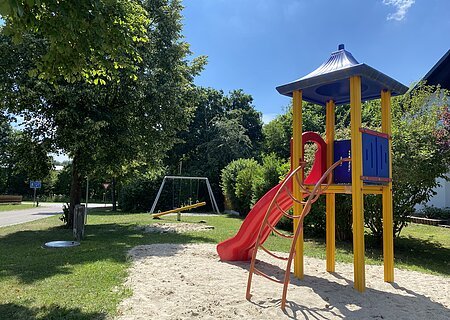 Spielplatz mit Rutsche, Klettergerüst und Schaukel in einem Wohngebiet auf Gras- und Sandfläche bei Sonnenschein.