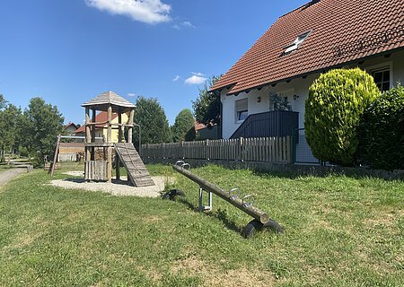 Spielplatz mit Wippe, Schaukel im Hintergrund und Kletterturm auf grünem Rasen, links vor einem Haus mit rotem Dach und Zaun an einem sonnigen Tag.