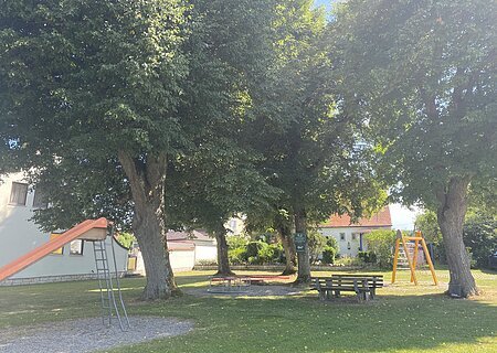 Spielplatz mit Rutsche, Karussell, Klettergerüst und Sitzbänken im Schatten unter großen Bäumen bei Wohnhäusern.