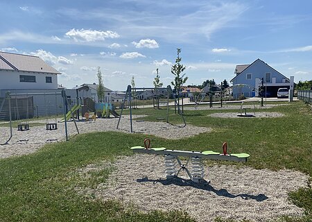 Moderner Spielplatz mit bunten Spielgeräten wie Wippe, Schaukeln, Klettergerüst und Rutsche umgeben von grüner Wiese und Schottersteinen in einer Wohnsiedlung bei sonnigem Himmel.