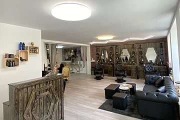 Friseursalon mit dunkelbraunen Holzmöbeln, schwarzen Friseurstühlen, schwarzem Sofa rechts im Bild und großen Spiegeln an der Wand. Links im Bild im Vordergrund eine Empfangstheke in Holzoptik auf hellem Bodenbelag.