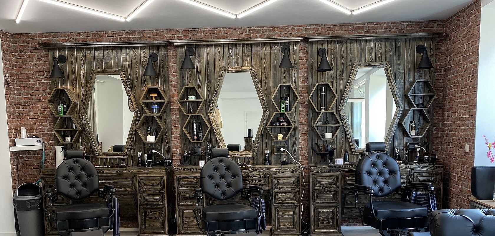 Friseursalon mit drei schwarzen Friseurstühlen vor dunkelbraunen Holzspiegeln und Backsteinwand. Rechts im Bild kleiner Ausschnitt vom Sitzbereich mit schwarzen Möbeln. An der Decke moderne Leuchte.