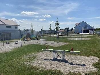 Moderner Spielplatz mit bunten Spielgeräten wie Wippe, Schaukeln, Klettergerüst und Rutsche umgeben von grüner Wiese und Schottersteinen in einer Wohnsiedlung bei sonnigem Himmel.