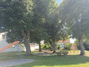 Spielplatz mit Rutsche, Karussell, Klettergerüst und Sitzbänken im Schatten unter großen Bäumen bei Wohnhäusern.