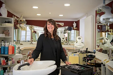 Frau steht lächelnd in einem Friseursalon neben einem Waschbecken und einem Wagen mit Friseurzubehör.