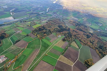 Luftaufnahme von Feldern, Waldflächen und einem Fluss bei Sonnenlicht, mit Dörfern und Straßen.