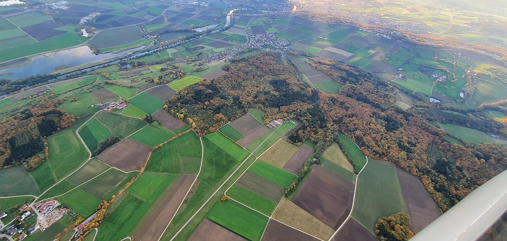 Luftaufnahme von Feldern, Waldflächen und einem Fluss bei Sonnenlicht, mit Dörfern und Straßen.